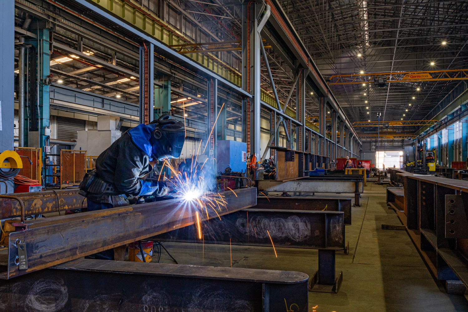 Image of someone in a welding suit welding a steel beam, there is sparks flying off the work area.